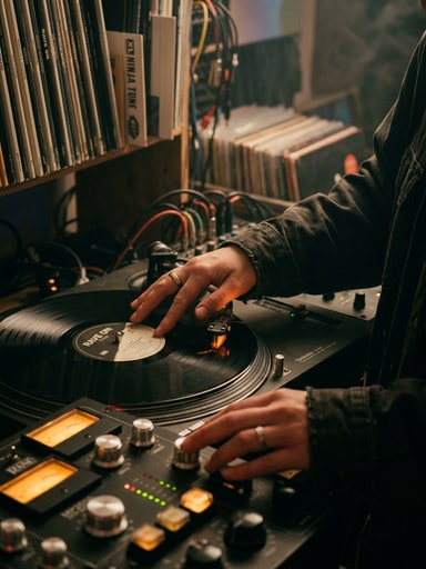Electronic artist cueing black vinyl records on a turntable beside an amber backlit analog mixer in a London studio for DJ-Kicks.
